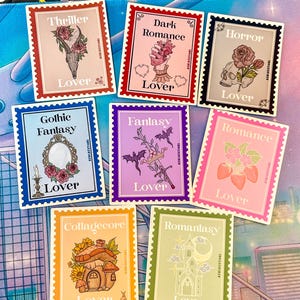 May include: A set of stickers in a postage stamp design, each with a different theme. Themes include "Thriller," "Dark Romance," "Horror," "Gothic Fantasy," "Fantasy," "Romance," "Collagecore," and "Romanlasy." The word "Lover" is printed on each sticker.