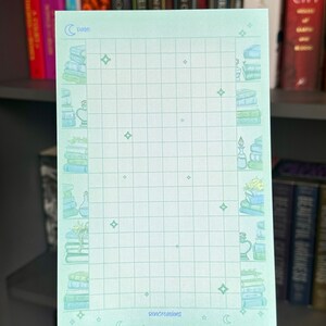 May include: A light blue gridded notepad with a whimsical book stack design on the left and right sides. The design features stacks of books, a bottle, and a plant. The notepad has a moon and stars design at the bottom and the text "Date" and "Rencreations" at the top.