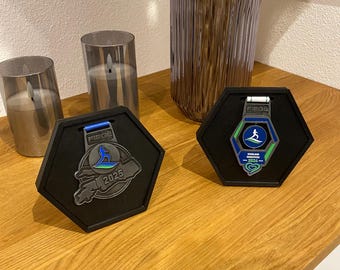 May include: Two hexagonal black display stands holding metallic marathon medals. The left medal features a blue runner graphic and the text FIEGL 2025. The right medal displays a blue runner graphic, the text FIEGL, MIDLAND MARATHON 2026, and a heart.