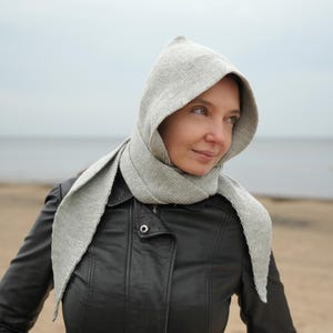 Könnte beinhalten: Ein hellgrauer Kapuzenschal mit langen, spitzen Enden. Der Schal ist aus einem Rippstrick gefertigt und wird über einer schwarzen Lederjacke getragen. Der Hintergrund zeigt einen Strand und einen bewölkten Himmel.