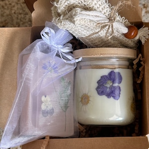 May include: A gift box filled with a candle, a small pouch with dried flowers, and a natural fiber washcloth. The candle is in a clear glass jar with a bamboo lid and features pressed flowers. The pouch is made of sheer white fabric and has a purple ribbon.