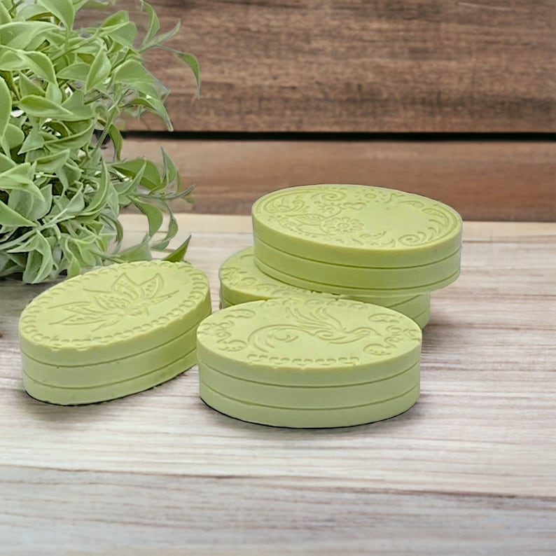 May include: Four oval-shaped green soap bars with decorative designs. The soaps are stacked on top of each other on a light wood surface.