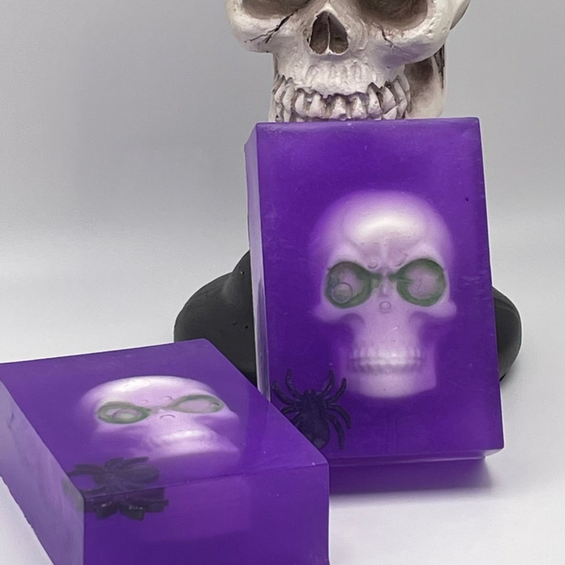 May include: Two purple soap bars with a white skull design and a black spider. The skull has green eyes.