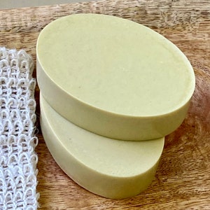 May include: Two oval-shaped bars of pale green soap stacked on top of each other. The soap is on a wooden surface.