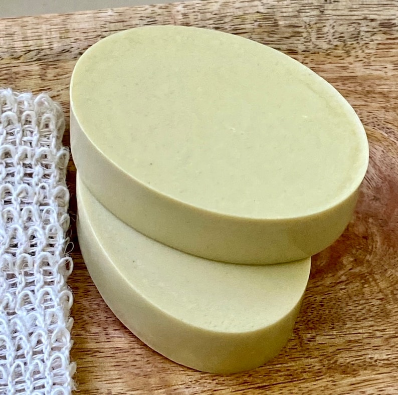 May include: Two oval-shaped bars of pale green soap stacked on top of each other. The soap is on a wooden surface.