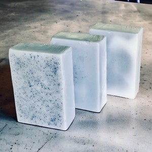May include: Three bars of soap, two are white and one is light blue with flecks of black. The bars are arranged in a row, with the blue bar on the left and the white bars on the right.