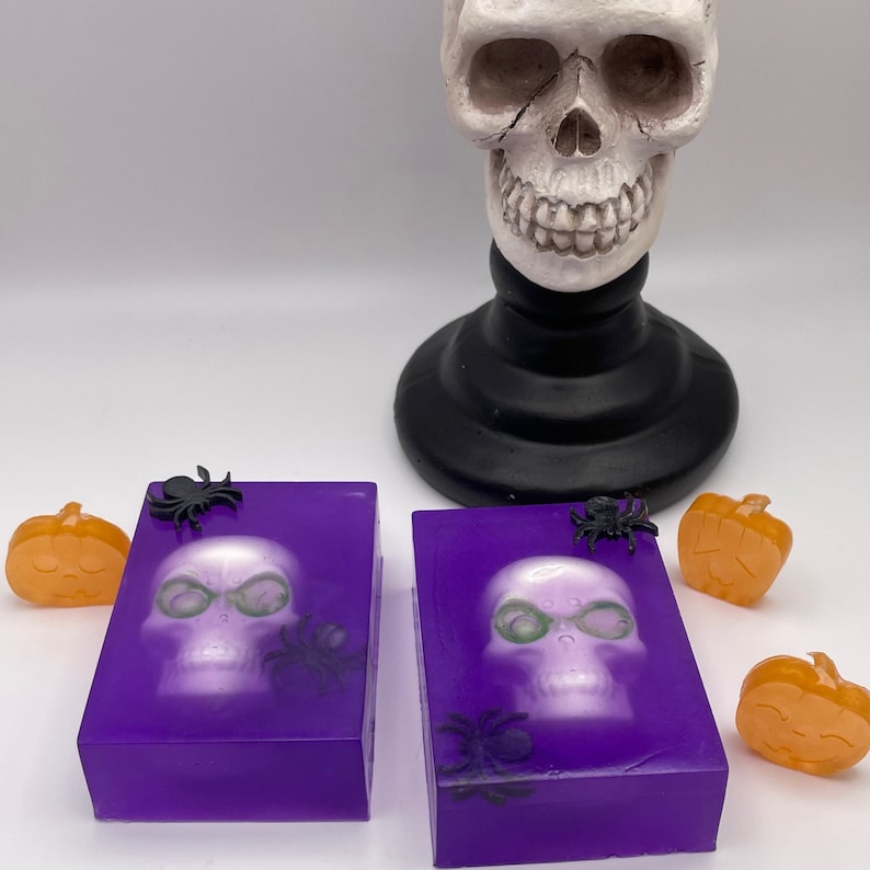 May include: Two purple soap bars with a white skull design and black spiders. The soap bars are on a white surface with orange pumpkin decorations.
