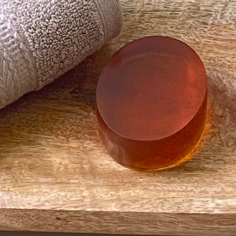 May include: A round, amber-colored soap bar on a wooden surface. The soap is smooth and shiny.
