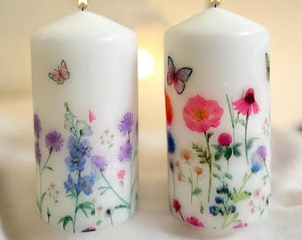 Può includere: Due candele bianche a forma di pilastro con disegni floreali. La candela a sinistra ha un disegno floreale blu e viola con una farfalla. La candela a destra ha un disegno floreale rosa e rosso con una farfalla.