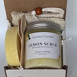 May include: A white gift box with a brown lid. The box contains a glass jar of lemon scrub, a wooden spoon, and a white washcloth. The jar is labeled "Signature Essential Lemon Scrub with Honey & Coconut Oil. All Natural | Handmade 8 oz | 227 g"
