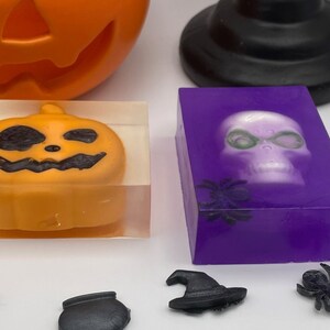 May include: Two Halloween themed soap bars. One bar is clear with a small orange pumpkin inside. The other bar is purple with a white skull inside. Both bars have black spiders on the front.