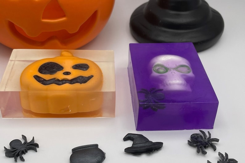 May include: Two Halloween themed soap bars. One bar is clear with a small orange pumpkin inside. The other bar is purple with a white skull inside. Both bars have black spiders on the front.