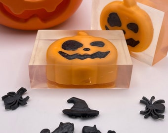 May include: Orange pumpkin-shaped soap bars with black facial features are encased in clear blocks. Black spider and witch shoe and hat decorations are scattered around the soap. An orange pumpkin sits in the background. Halloween-themed.