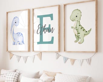 May include: Three framed watercolor dinosaur prints. One features a blue Brontosaurus, another a teal letter "E" with the name "Ethan", and the third a green T-Rex. A garland of triangle flags hangs below the frames.