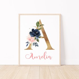 May include: Framed art print featuring a gold glitter letter "A" adorned with watercolor flowers in navy, blush pink, and green. The name "Amelia" is written in pink script below the letter. The artwork is displayed in a light wood frame.