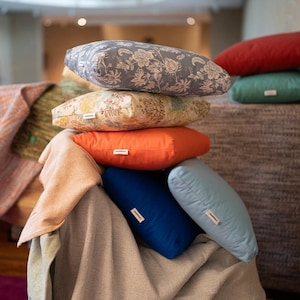Könnte beinhalten: Ein Stapel bunter Stützkissen in verschiedenen Farben und Mustern. Die Kissen umfassen Blumendrucke, einfarbiges Orange, Blau und Hellblau. Jedes Kissen hat ein rechteckiges Etikett. Die Kissen sind auf einer neutralfarbenen Decke gestapelt.