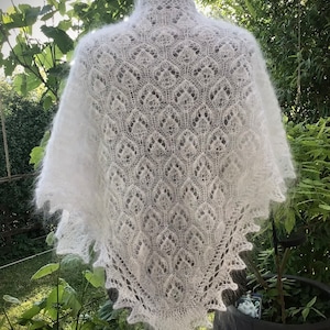 Op de afbeelding: Een witte kanten sjaal met een delicaat patroon, getoond op een wit mannequin tegen een groene, bladerrijke achtergrond.