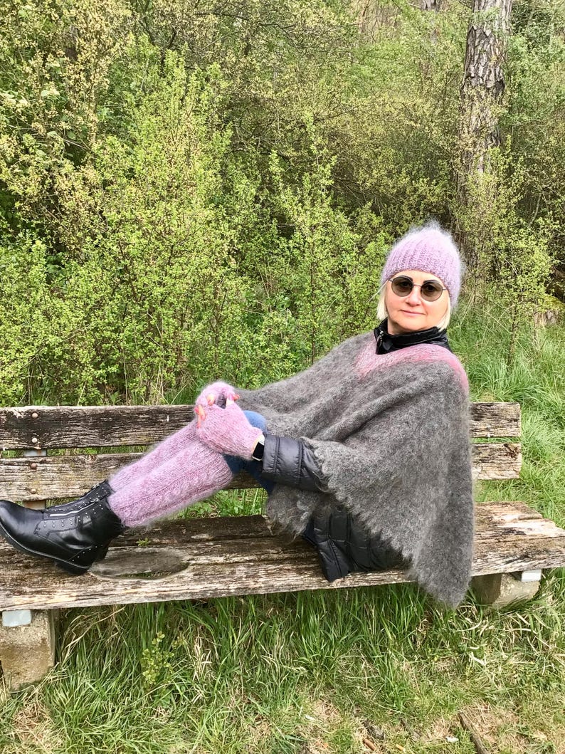 May include: A woman wearing a gray and black poncho, a pink fuzzy hat, and pink fuzzy leg warmers. She is sitting on a wooden bench in a forest setting.