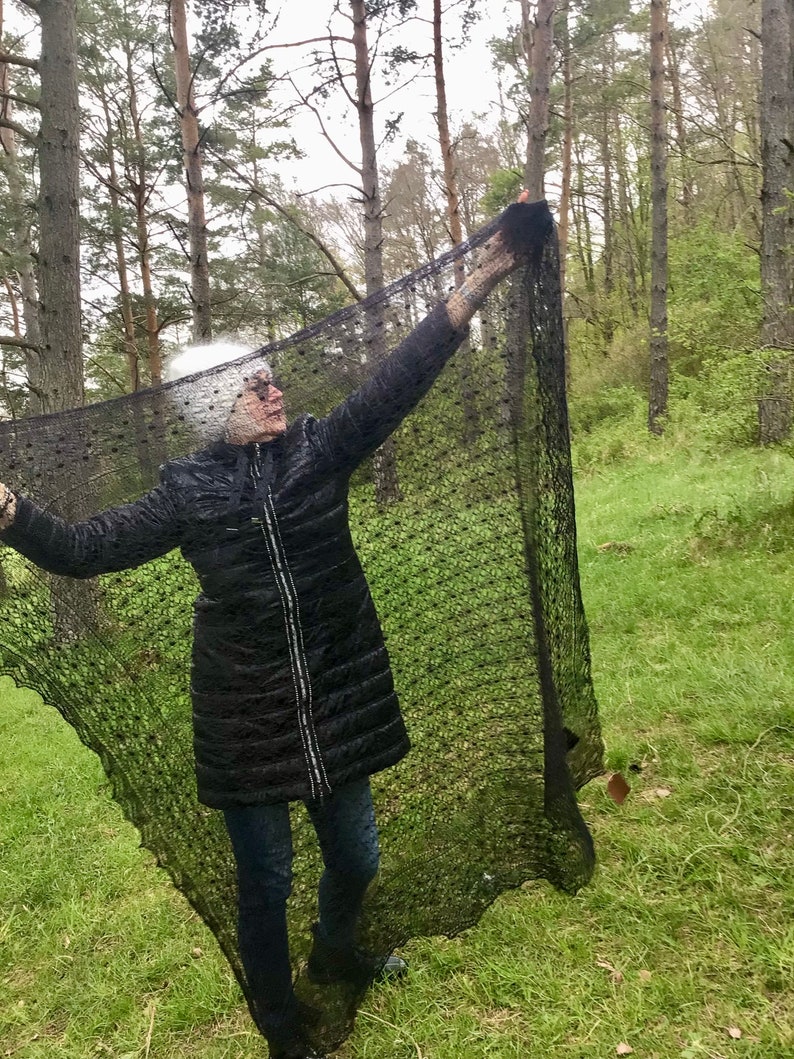 Puede incluir: Una persona con una chaqueta negra y pantalones vaqueros azules sostiene un chal de encaje negro con un patr&oacute;n de puntos. El chal est&aacute; drapeado sobre el cuerpo de la persona y se sostiene con los brazos. La persona est&aacute; de pie en un entorno boscoso.