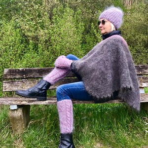 May include: A woman wearing a purple knitted hat, a gray poncho, and purple leg warmers sits on a wooden bench. She is wearing black boots and blue jeans.