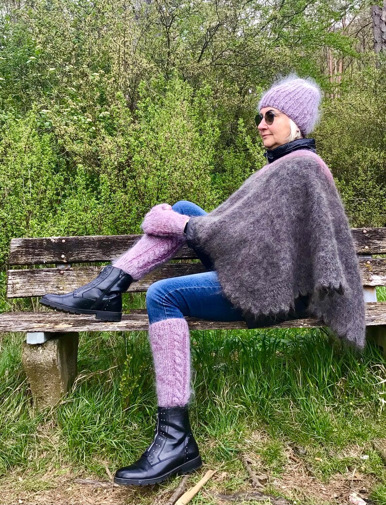 May include: A woman wearing a purple knitted hat, a gray poncho, and purple leg warmers sits on a wooden bench. She is wearing black boots and blue jeans.