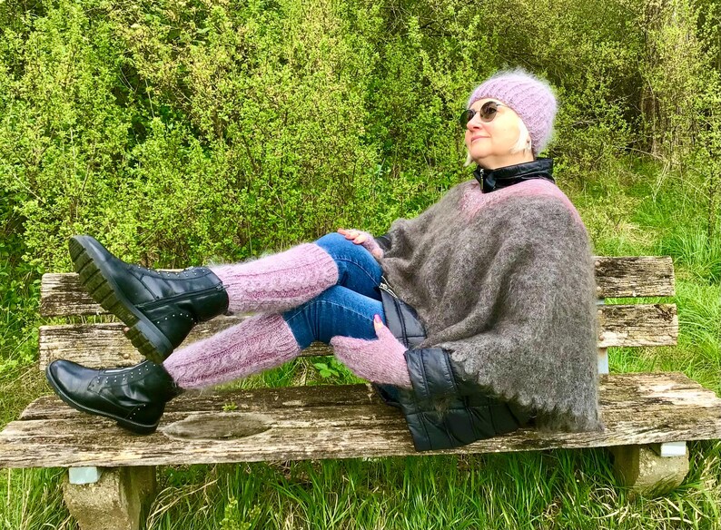 May include: A woman wearing a pink knit hat, sunglasses, a brown fuzzy poncho, and black boots sits on a wooden bench. She is wearing jeans and pink fuzzy socks.