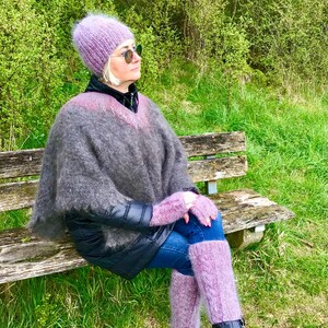 May include: A woman wearing a purple knitted hat, a gray and purple poncho, purple leg warmers, and black boots sits on a wooden bench in a grassy area.