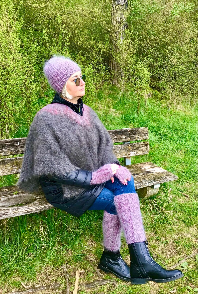 May include: A woman wearing a purple knitted hat, a gray and purple poncho, purple leg warmers, and black boots sits on a wooden bench in a grassy area.