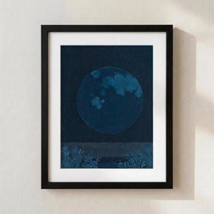 May include: A framed wall art print featuring a large, textured blue moon centred in a dark navy night sky above a dark horizon line with silhouettes of low-lying vegetation at the bottom, all encased in a simple black frame with a white mount.
