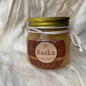 May include: A glass jar of honey with a gold lid and a brown label that says "Baska Ethiopian Highland White Honey".