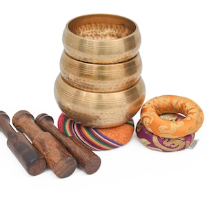May include: A stack of three brass singing bowls with a hammered texture, resting on colorful cushions. Four wooden mallets are arranged in front. Two additional cushions are also present, one orange and one purple.