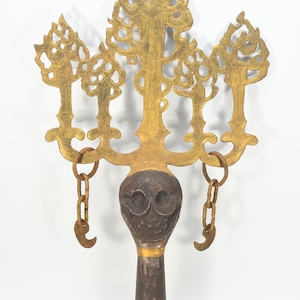 Puede incluir: Objeto metálico antiguo y ornamentado, con una parte superior dorada en forma de árbol y una base oscura en forma de calavera. El objeto tiene un mango marrón oscuro con detalles dorados y pequeñas cadenas con ganchos.