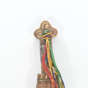 May include: The top of a decorative sword handle with a gold and silver finish. The handle is adorned with a multi-colored braided cord.