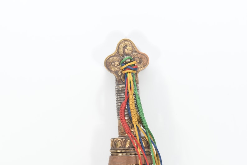 May include: The top of a decorative sword handle with a gold and silver finish. The handle is adorned with a multi-colored braided cord.