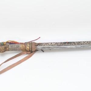 May include: A silver and gold decorated dagger with a leather strap and colorful tassels. The dagger has intricate floral designs and a sharp blade.