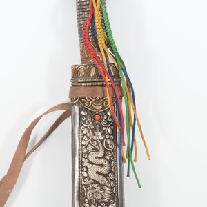 May include: A silver and gold decorated scabbard with a leather strap and colorful tassels. The scabbard has a dragon design and a red stone accent.
