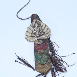 May include: A decorative pendant featuring a seated Buddha figure within a leaf-shaped frame. The pendant is attached to a colorful fabric strip with tassels and a brown cord for hanging. The design incorporates gold, red, green, and brown hues.