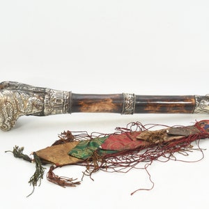 May include: A long, brown wooden ceremonial horn with silver embellishments and a decorative tassel. The horn is decorated with intricate carvings and has a silver tip. The tassel is made of red, green, and gold fabric.