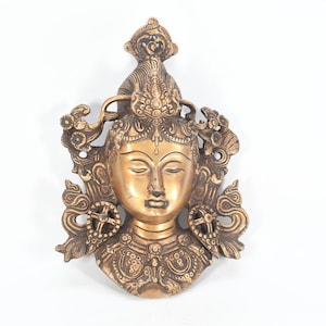 May include: A gold-colored metal wall hanging depicting a deity with a serene expression. The deity is adorned with intricate details and a crown-like headdress.
