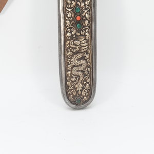 May include: A silver and gold scabbard with a dragon design and green and red gemstones. The scabbard is decorated with intricate floral patterns.