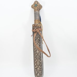 May include: A silver and gold decorated dagger with a brown leather strap. The handle is ornate and features a turquoise stone.