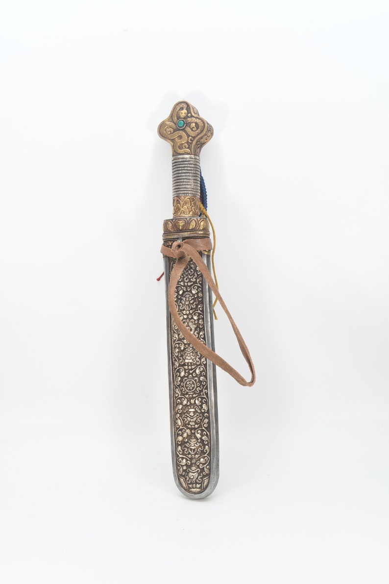 May include: A silver and gold decorated dagger with a brown leather strap. The handle is ornate and features a turquoise stone.