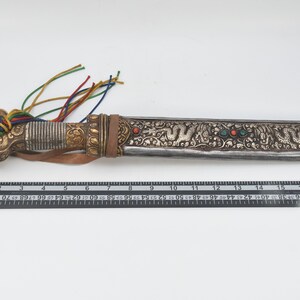 May include: A silver and gold ceremonial dagger with a leather handle and a decorative tassel. The blade is decorated with intricate floral and dragon designs and features red and green gemstones.