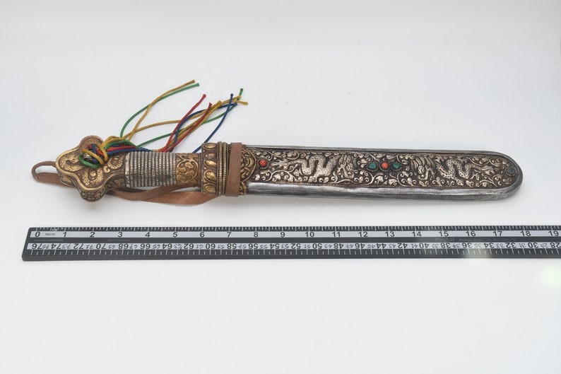 May include: A silver and gold ceremonial dagger with a leather handle and a decorative tassel. The blade is decorated with intricate floral and dragon designs and features red and green gemstones.