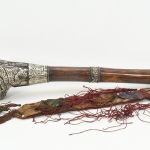 May include: A long, brown wooden ceremonial horn with silver embellishments and a ram's head at the end. The horn is decorated with intricate carvings and has a red and green tassel attached.