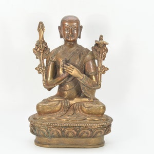 Puede incluir: Una estatua de metal dorado de una figura de Buda sentada con una expresión serena. El Buda está adornado con detalles intrincados y sostiene un gesto de mano frente a su pecho. La estatua se coloca sobre una base decorativa.