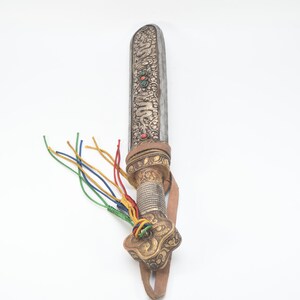 May include: A silver and gold ceremonial dagger with a leather strap and colorful tassels. The blade is decorated with intricate carvings and gemstones.
