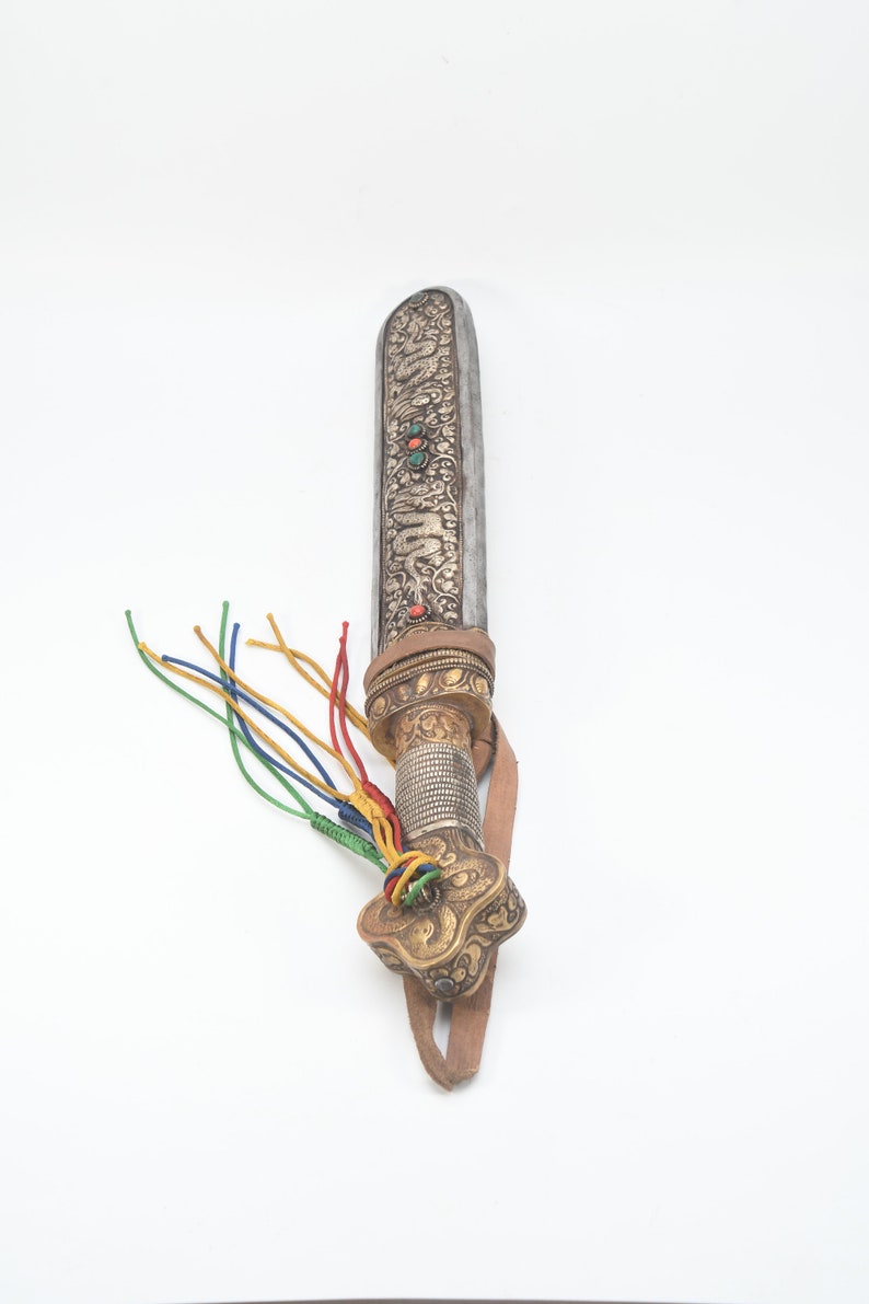 May include: A silver and gold ceremonial dagger with a leather strap and colorful tassels. The blade is decorated with intricate carvings and gemstones.