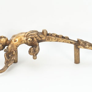 May include: A gold-toned metal door handle shaped like a cherub with wings. The cherub is lying on its back with its head turned to the side. The handle is attached to a metal rod.