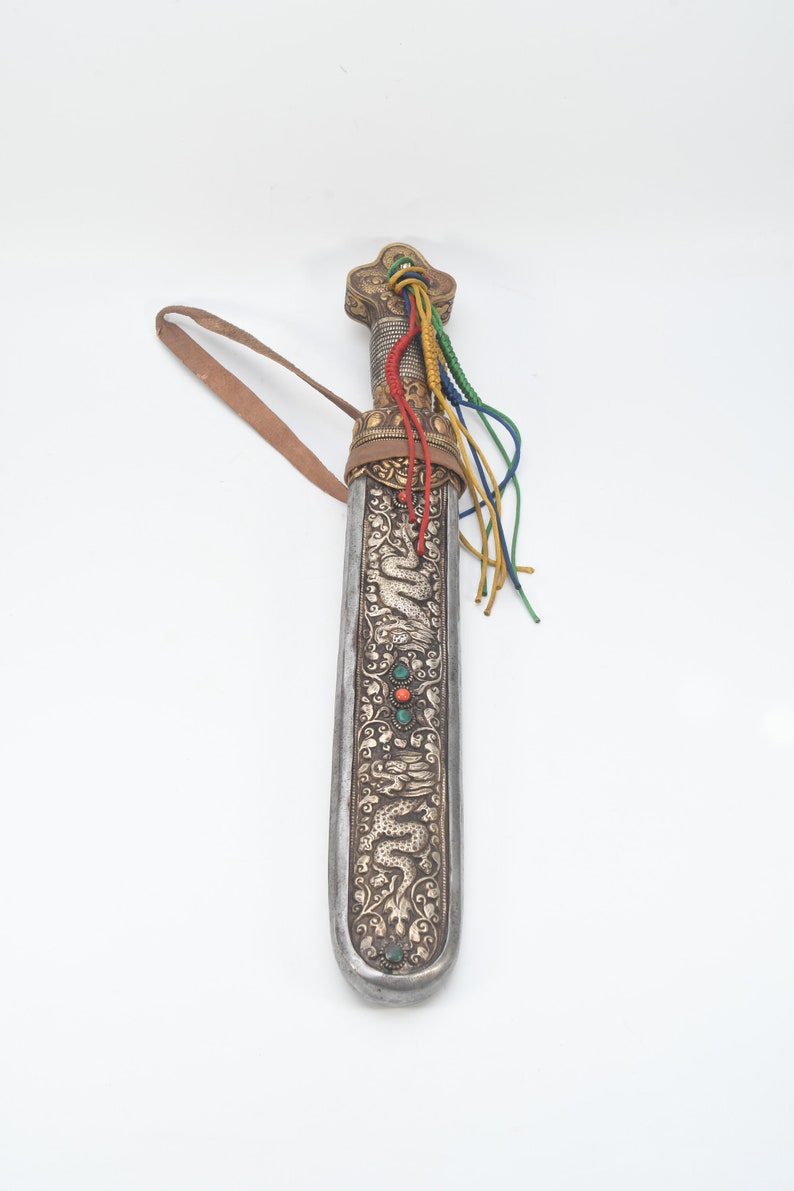 May include: A silver and gold scabbard with intricate dragon and floral designs. The scabbard has a leather strap and a colorful tassel.